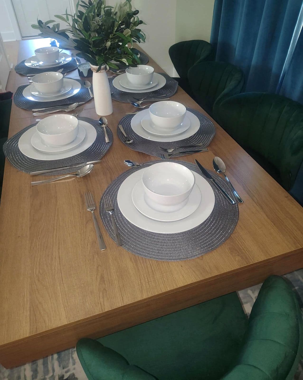 Dining table set with white dishes on gray chargers beside green velvet chairs